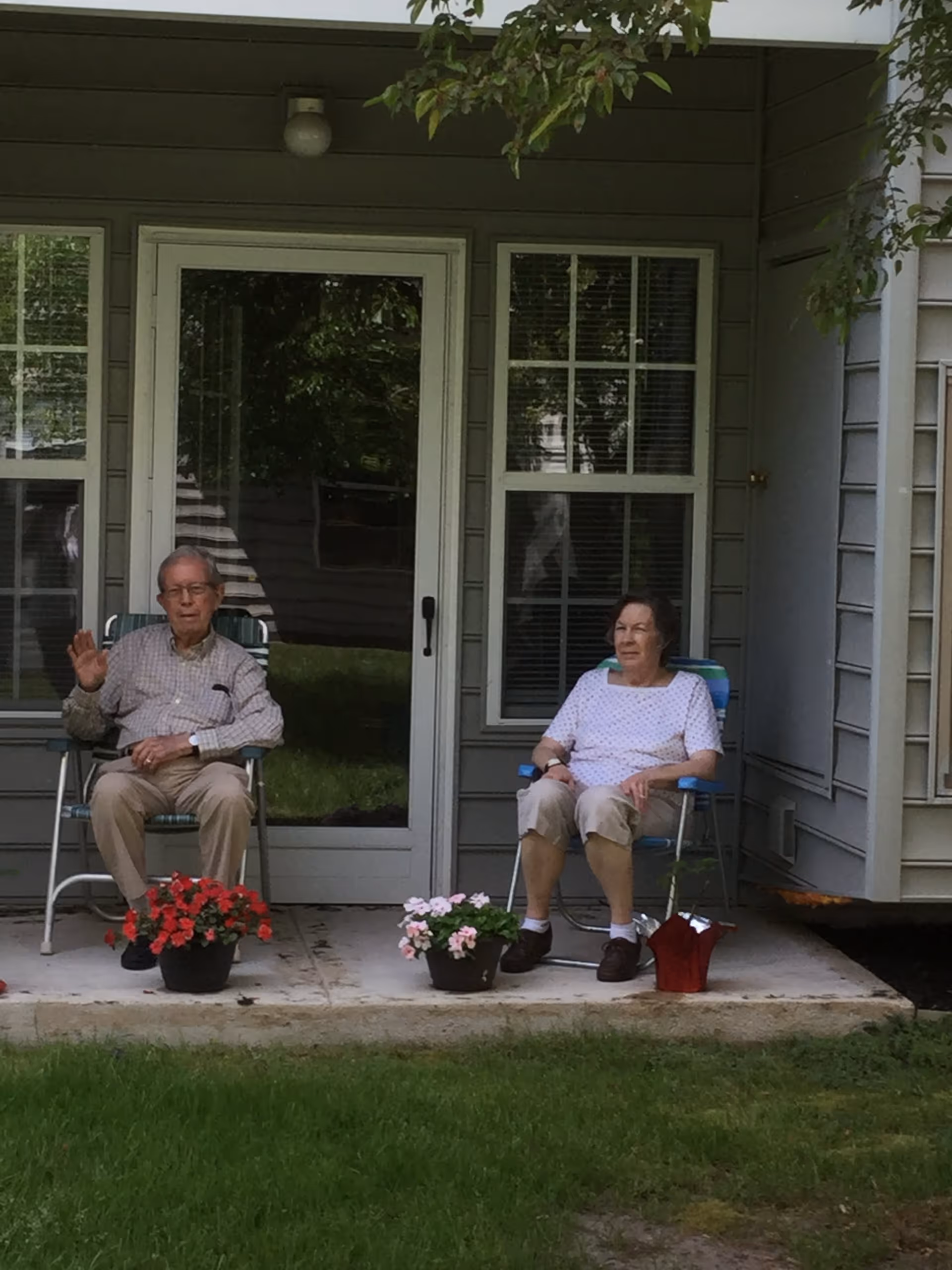 Two elderly individuals sitting on lawn chairs on a small concrete patio outside a house. There are potted flowers placed on the patio in front of them. The house has a glass door and windows with blinds, and some greenery is visible in the background.