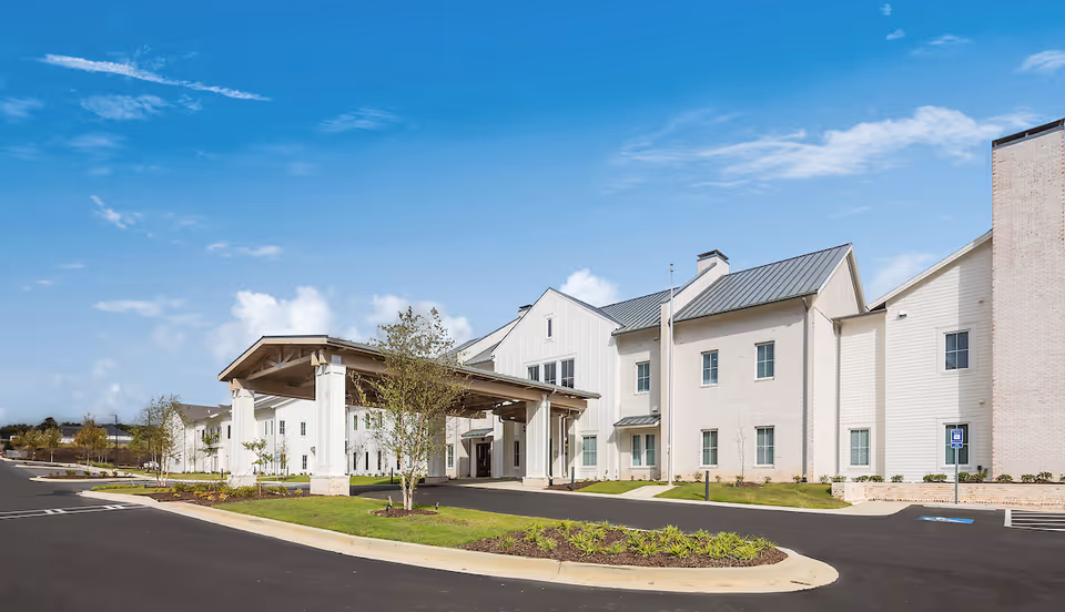 Exterior view of The Claiborne at Shoe Creek senior living facility showing a large covered entrance with white pillars, a paved driveway, landscaped greenery, and a clear blue sky with some clouds.