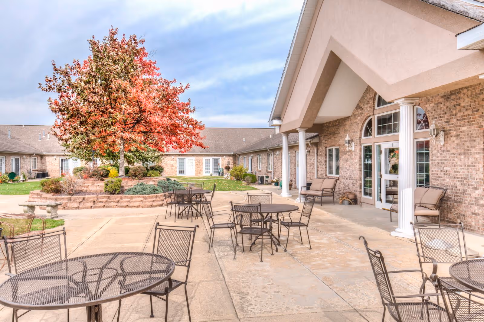 Outdoor patio area at Brookstone Estates of Effingham featuring several metal tables and chairs on a concrete surface, a large tree with red and green leaves in the background, and a brick building with white columns and cushioned seating along the wall.