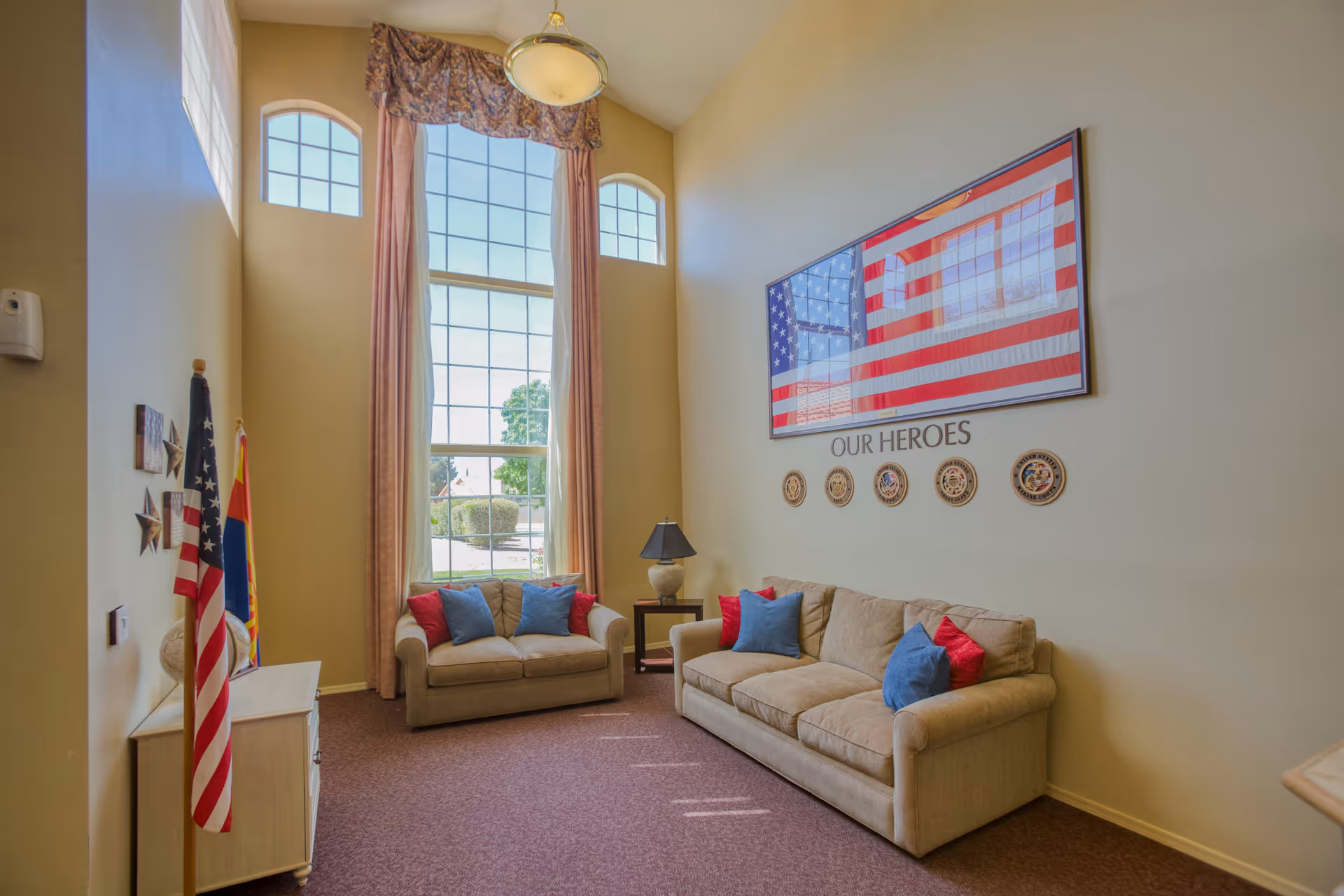 A living room area with two beige sofas adorned with red and blue pillows. A large window with three sections, including two arched windows at the top, lets in natural light. The wall features a framed American flag with the words 'OUR HEROES' and military emblems beneath it. There are two flags standing in the corner and a small side table with a lamp between the sofas.