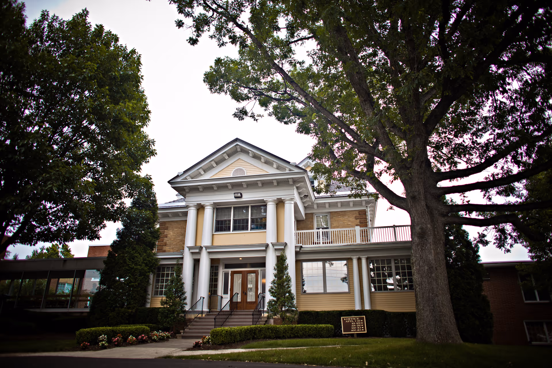 Front exterior view of a senior living community building with large white columns, a staircase leading to double wooden doors, surrounded by trees and well-maintained landscaping.