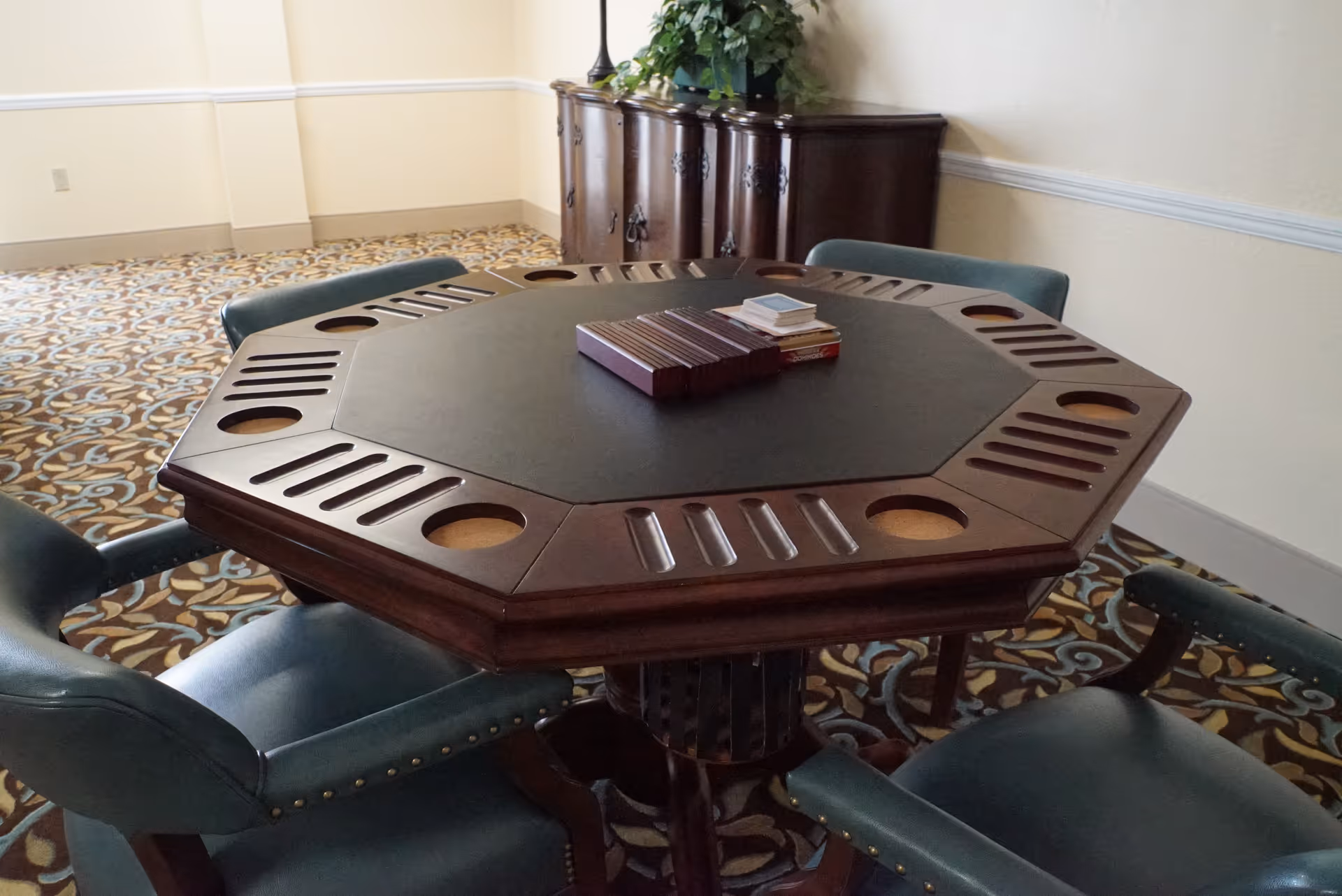 Octagonal wooden game table with slots and cup holders, surrounded by four green upholstered chairs, set on a patterned carpeted floor in a room with cream-colored walls and a wooden cabinet with a plant on top in the background.