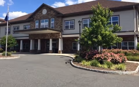 Front exterior view of CareOne at Somerset Valley Assisted Living facility showing a two-story building with a covered entrance, multiple windows, a flagpole, and landscaped greenery including bushes and a tree.