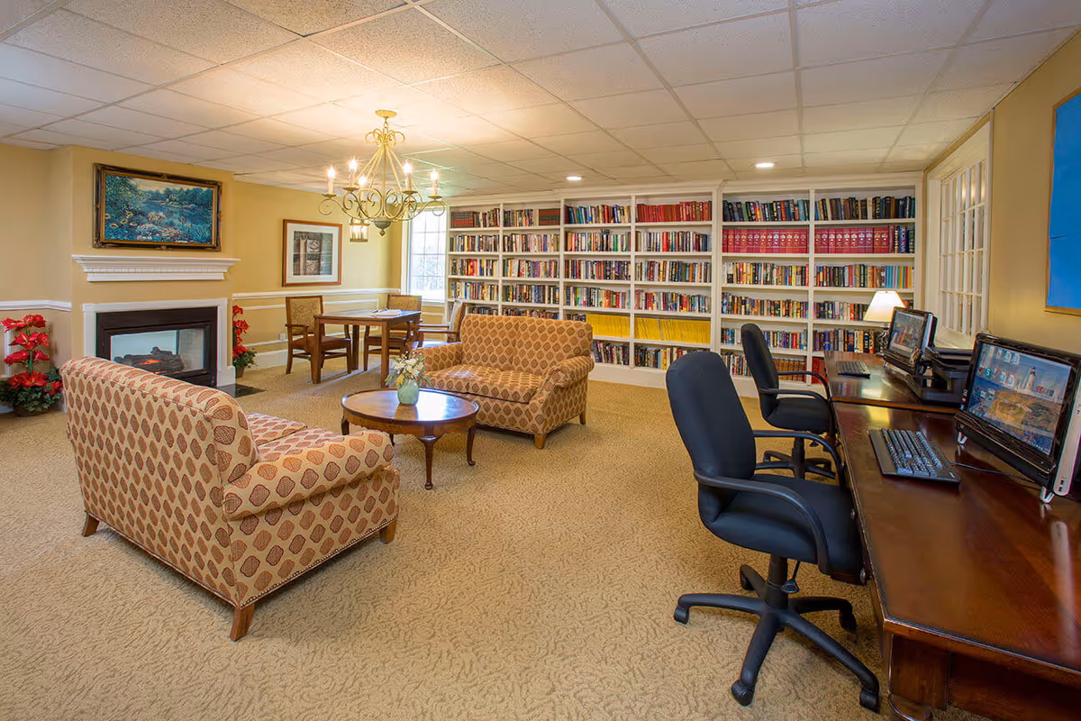 A cozy common room with two patterned sofas facing each other around a wooden coffee table with a small flower vase. Behind the sofas is a fireplace with a painting above it and red flowers on either side. To the right, there are two computer workstations with black office chairs and desktop computers. The back wall features large white bookshelves filled with books. A chandelier hangs from the ceiling, and there is a small table with chairs near a window in the background.