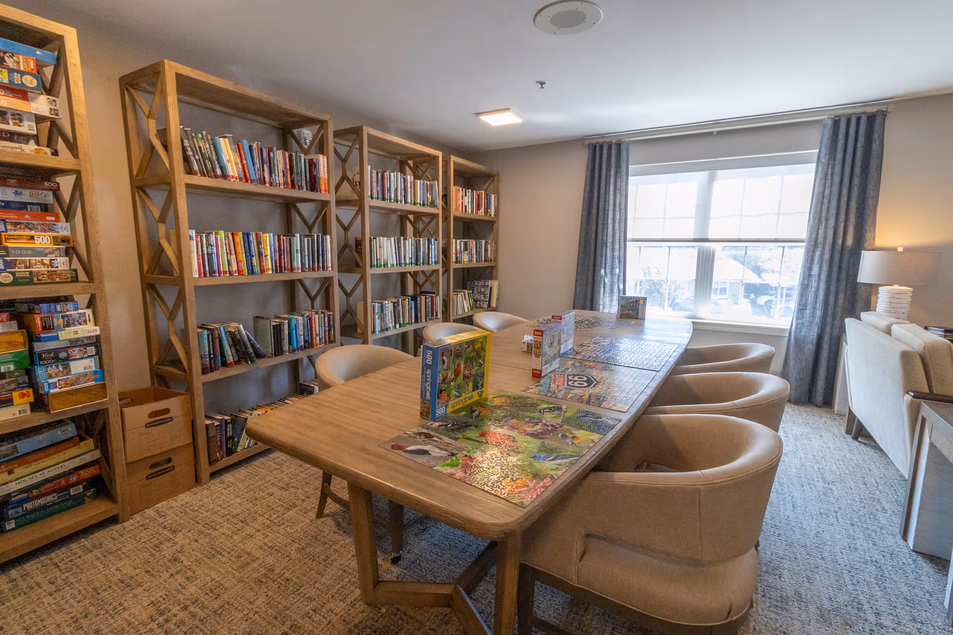 A cozy room with a large wooden table surrounded by beige upholstered chairs. Several jigsaw puzzles are laid out on the table, some partially completed. Behind the table, there are wooden bookshelves filled with books and board games. A large window with gray curtains lets in natural light, and a lamp is visible on a side table near a beige sofa.