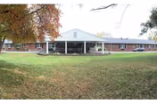 Front exterior view of a single-story brick building with a white covered entrance and a large grassy lawn in front, surrounded by trees with autumn foliage.
