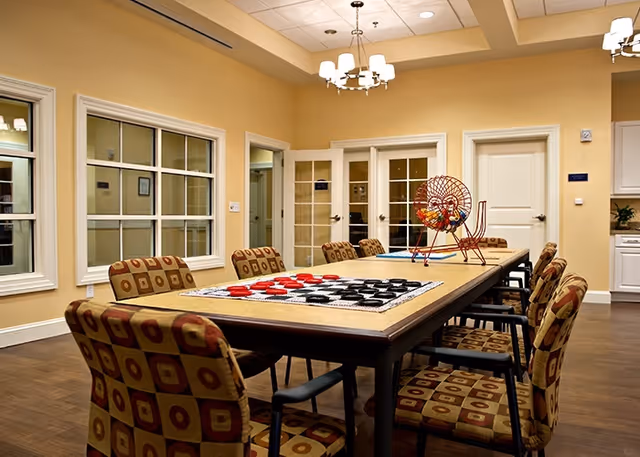 A well-lit room with a long table surrounded by patterned cushioned chairs. On the table, there is a large checkers game set up and a bingo cage with numbered balls. The room has beige walls, a chandelier, and multiple doors and windows.