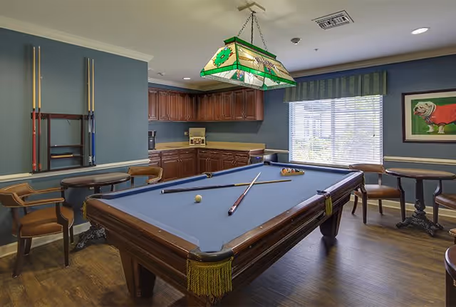 A recreational room with a pool table in the center, pool cues mounted on the wall, wooden chairs and small round tables around the room, a window with blinds and a green-striped valance, wooden cabinets along the back wall, and a stained glass hanging light fixture above the pool table.