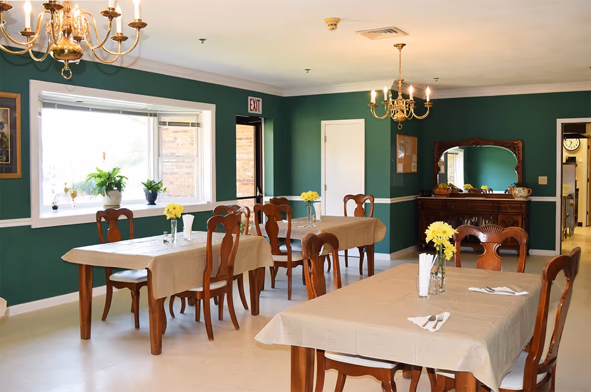 A dining room with green walls and white trim, featuring wooden tables covered with beige tablecloths and wooden chairs. Each table has a small vase with yellow flowers and utensils. There are two chandeliers hanging from the ceiling, a window with plants on the sill, an exit door, and a wooden sideboard with a large mirror on the wall.