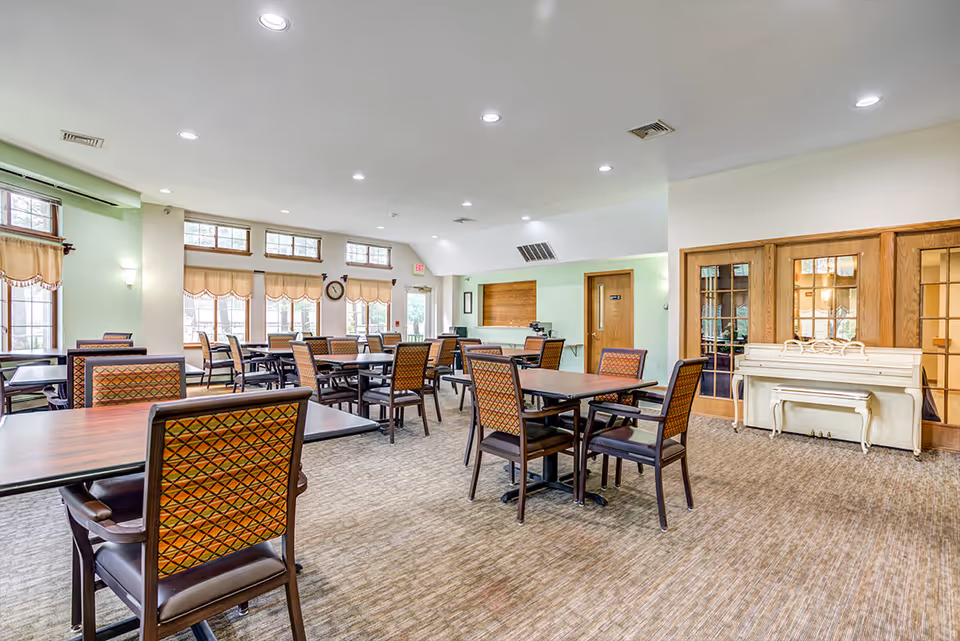 A spacious dining room with multiple wooden tables and patterned chairs arranged neatly. The room has large windows with beige curtains allowing natural light to fill the space. There is a white upright piano against a wall with wooden framed glass doors nearby. The walls are painted light green and white, and the ceiling has recessed lighting.