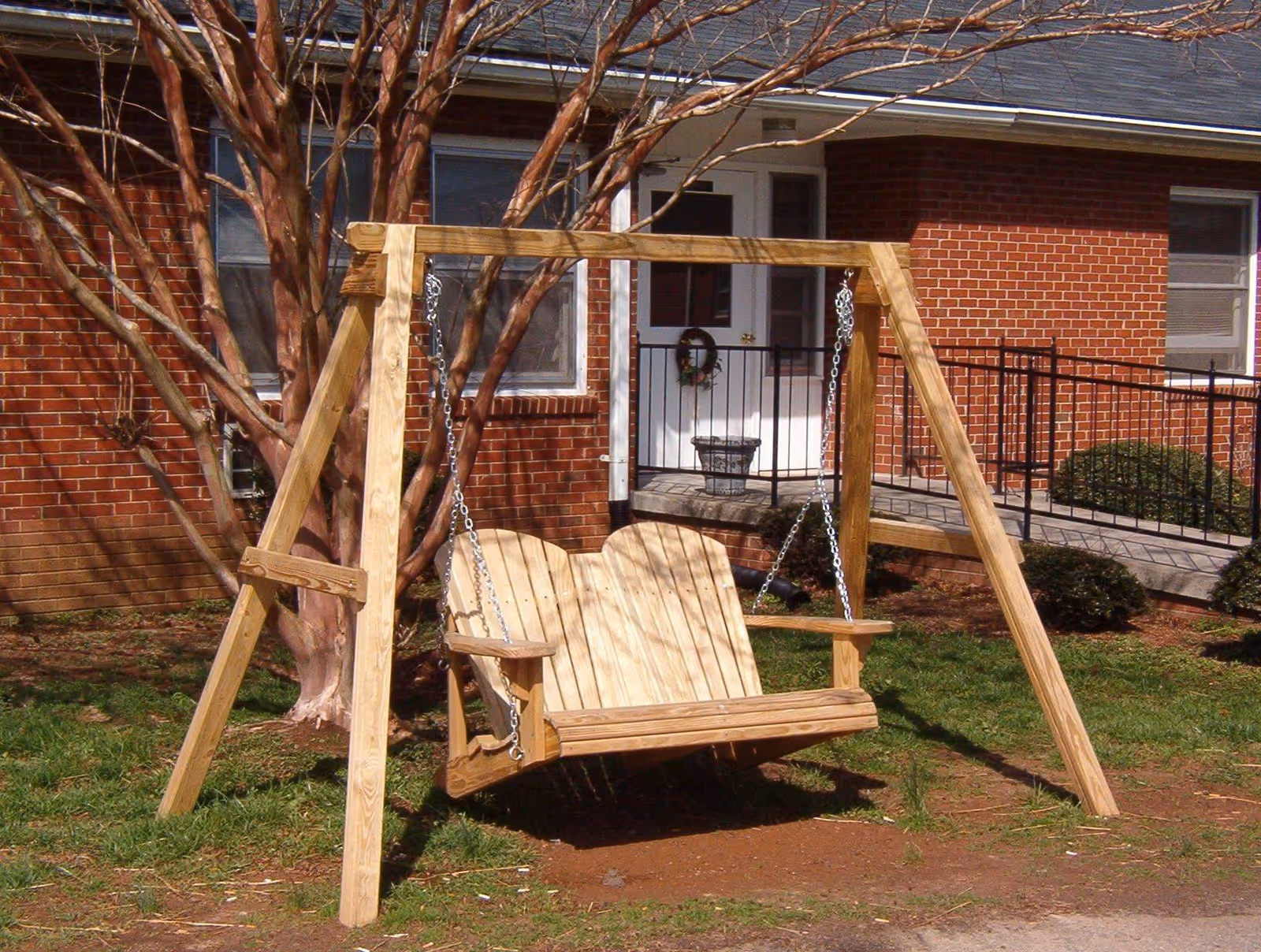 A wooden outdoor swing with a heart-shaped backrest hanging from a wooden frame on a grassy area in front of a brick building with windows and a door.