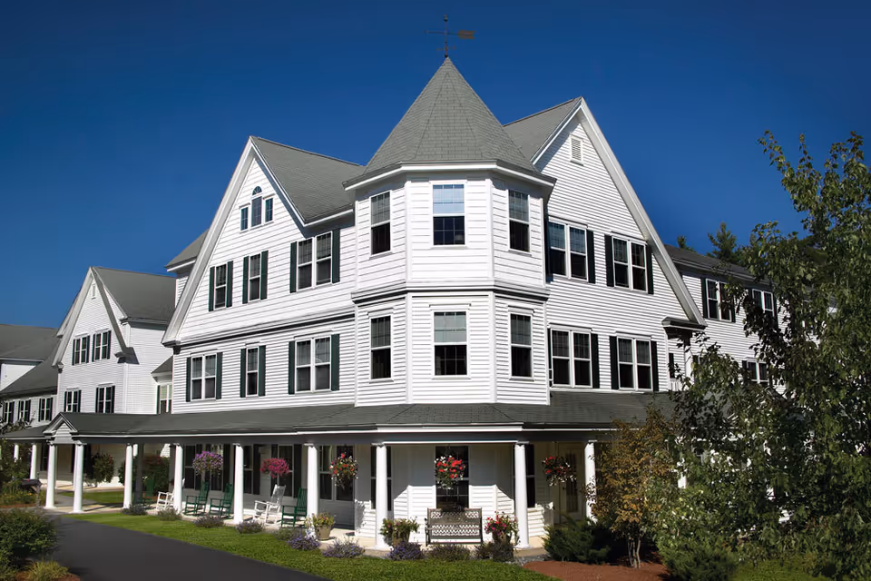 Front exterior of a large white multi-story building with a wraparound porch, columns, rocking chairs, and hanging flower baskets.