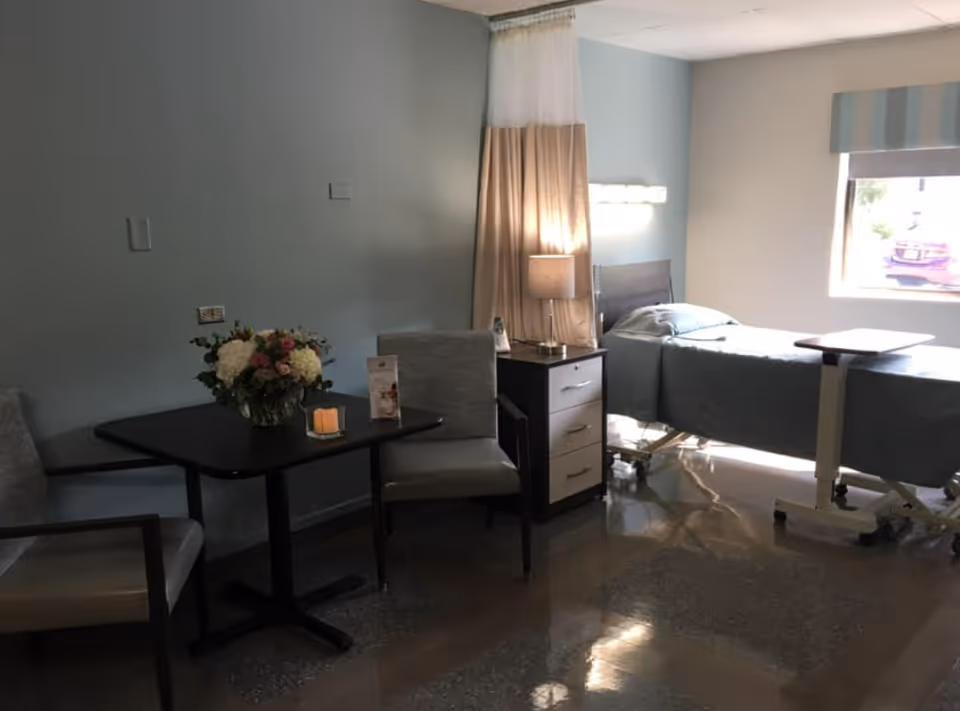 A room with a hospital bed next to a window with striped curtains. There is a bedside table with a lamp and a remote control. In the foreground, there is a small table with a flower arrangement and two chairs. The floor is shiny and reflective.