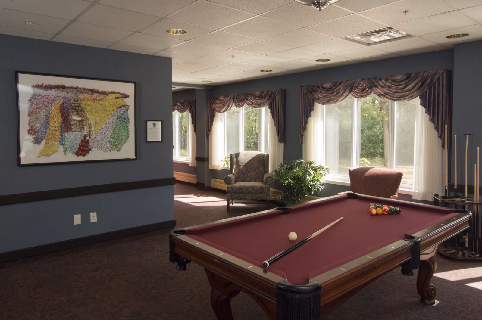 A recreational room with a burgundy pool table set up for a game, including a cue stick and billiard balls. The room has large windows with striped valances and sheer curtains, letting in natural light. There are two upholstered armchairs near the windows and a green potted plant. The walls are painted blue with a colorful abstract painting hanging on one wall.