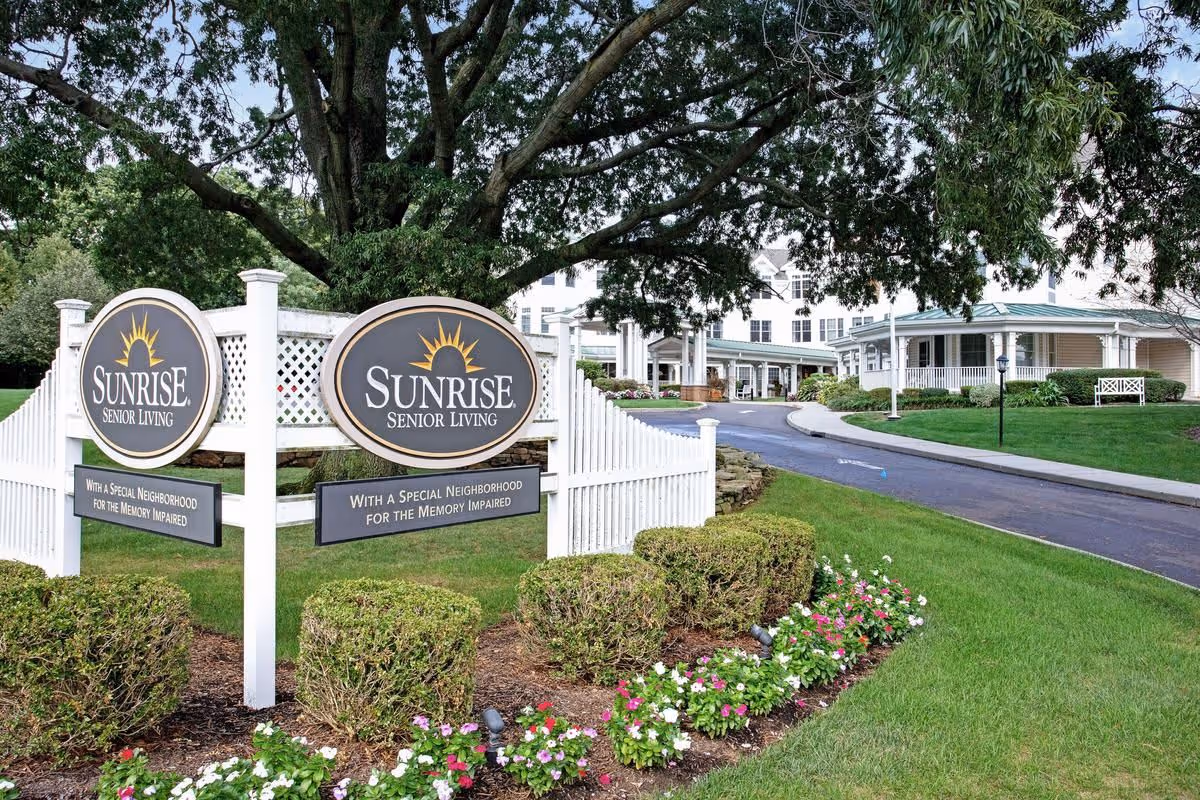 Entrance to Sunrise Senior Living facility with two large signs displaying the name and tagline 'With a Special Neighborhood for the Memory Impaired,' surrounded by manicured bushes, flowers, a white fence, and a large tree, with the building and driveway visible in the background.