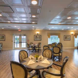 A spacious dining room with multiple round tables set with folded white napkins and small flower centerpieces. The room features wooden flooring, beige walls, framed artwork, large windows with yellow valances, and a decorative ceiling with recessed lighting. Double glass doors are visible at the far end of the room.