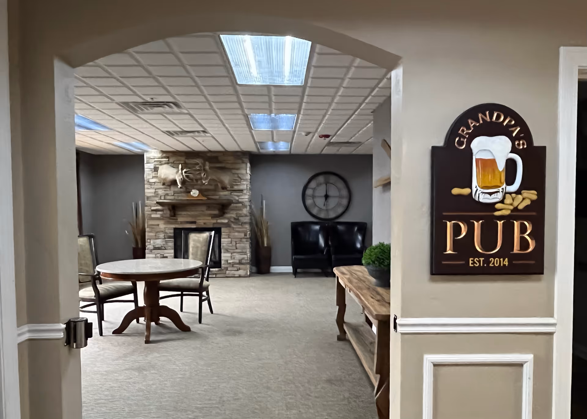 View through an arch into a communal lounge with a round table and chairs, a stone fireplace, seating, and a "Grandpa's Pub" sign on the wall.