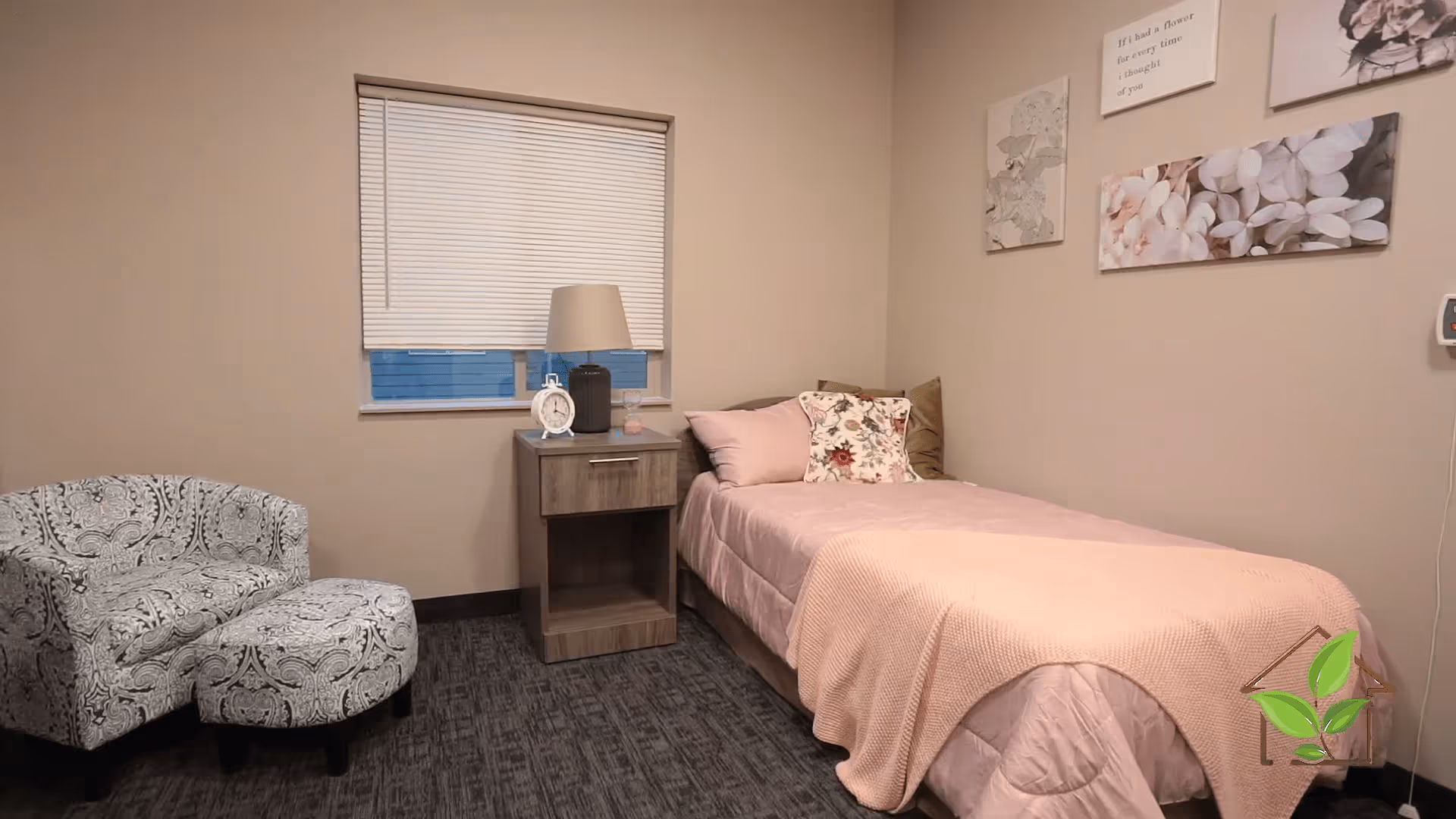 A small bedroom with a single bed covered in a pink blanket and floral pillows. Next to the bed is a wooden nightstand with a lamp, a clock, and a small decorative item. A patterned armchair with a matching ottoman is placed near the window with closed blinds. The walls are decorated with floral and inspirational artwork.