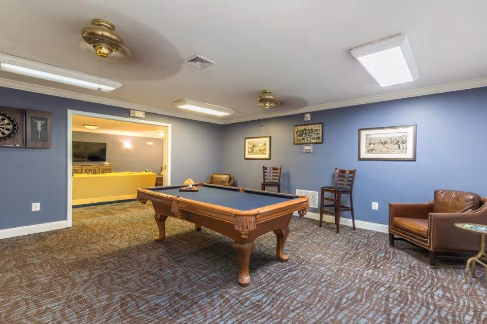 A recreational room with a pool table in the center, blue walls, and carpeted floor. The room has two ceiling fans with lights, framed pictures on the walls, two wooden chairs, and a leather armchair with a small side table. An open doorway leads to another room with a yellow couch and a TV.