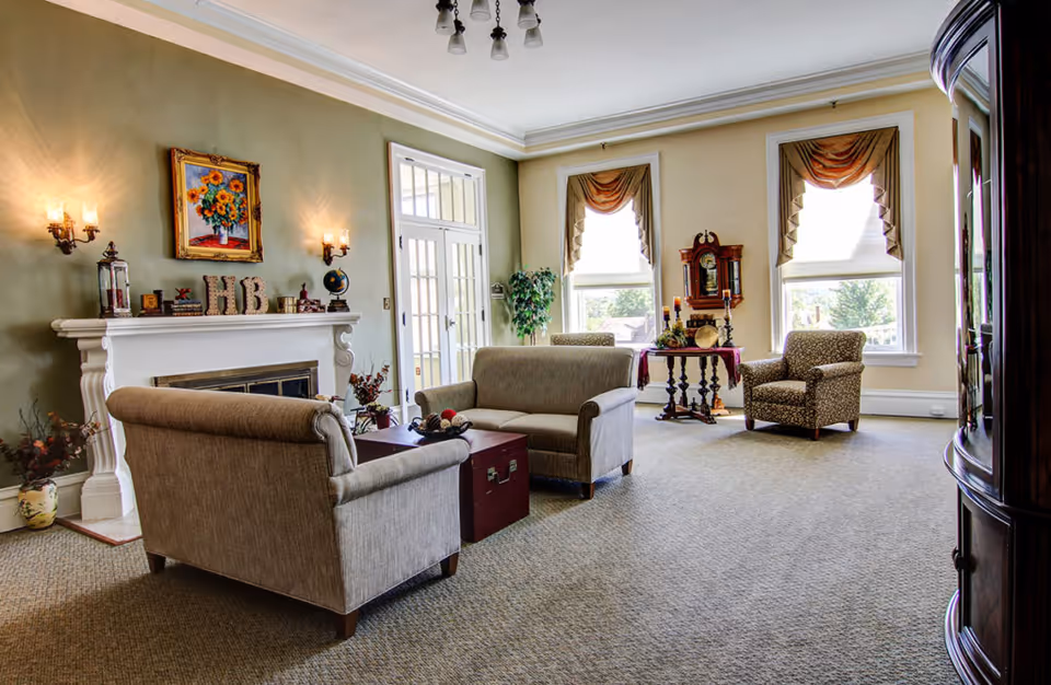A cozy senior living community lounge with beige armchairs and a loveseat arranged around a small wooden chest used as a coffee table. The room features a white fireplace with decorative items and a painting of sunflowers above it. Two large windows with draped curtains let in natural light, and a wooden grandfather clock and a small table with candles and decor are positioned near the windows. The walls are painted in soft green and cream tones, and the carpet is light brown.