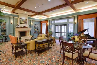 Elegant senior living lounge with seating, fireplace, grand piano, and patterned carpet.