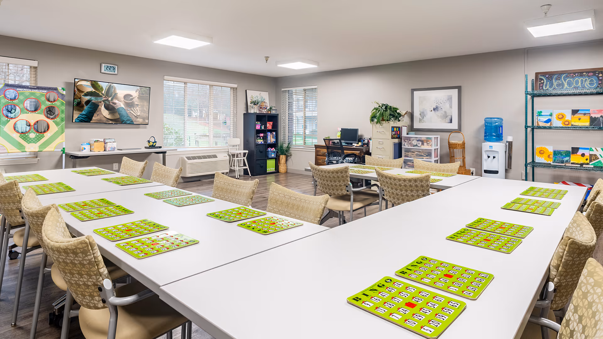 A bright and spacious activity room with tables arranged in a U-shape, each table set with multiple green bingo cards. The room has several beige patterned chairs around the tables, large windows letting in natural light, a water cooler, shelves with colorful paintings, a welcome sign, and a desk with a computer in the background.