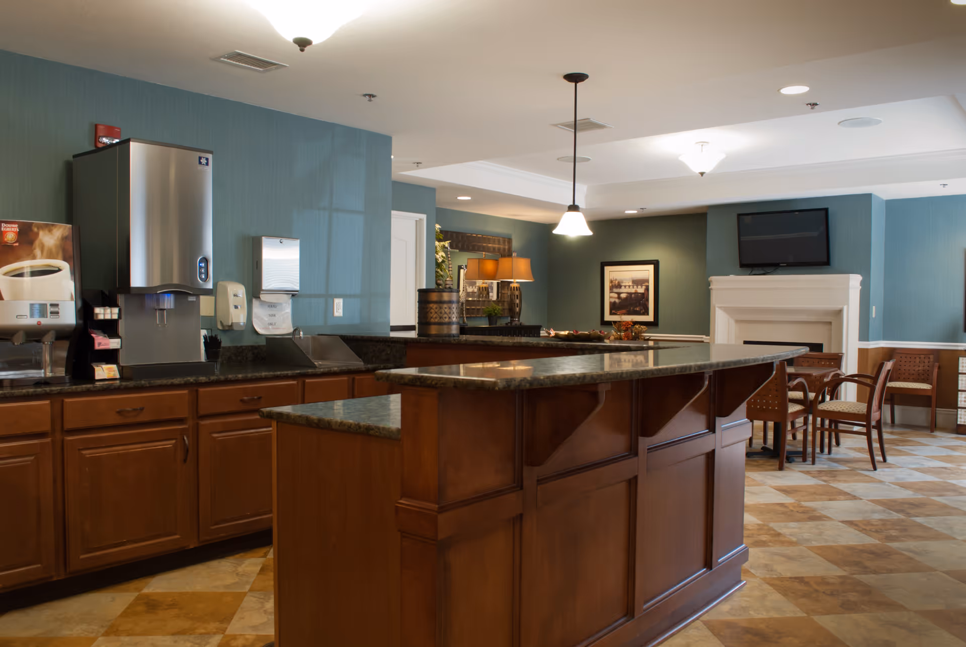 Communal dining/lounge area with a large wooden service counter, coffee machines, seating, a TV above a fireplace, and pendant lighting.