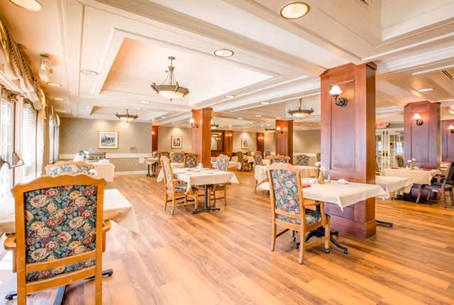 A spacious dining room with wooden floors, multiple tables covered with white tablecloths, and chairs with floral upholstery. The room features warm lighting, wooden columns, framed artwork on the walls, and large windows with curtains allowing natural light to enter.