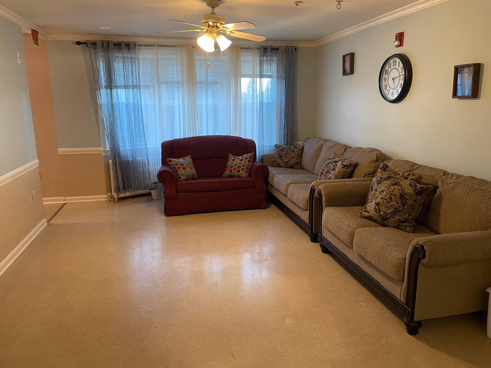 A living room with a beige couch and loveseat, a red loveseat with patterned pillows, a ceiling fan with lights, a large window with sheer curtains, and a wall clock above the couch.