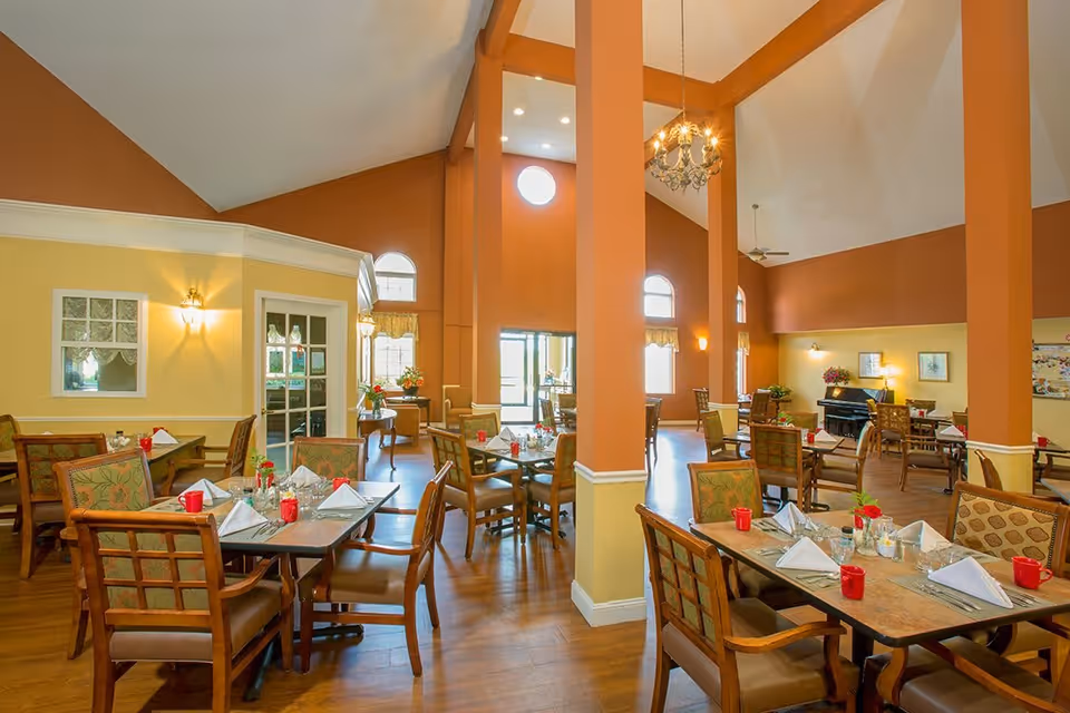 A spacious dining room with high vaulted ceilings and large orange pillars. Multiple tables are set with white napkins, red cups, and silverware. The room has warm yellow and orange walls, wooden floors, and several windows letting in natural light. There is a piano in the back corner and various chairs arranged around the tables.