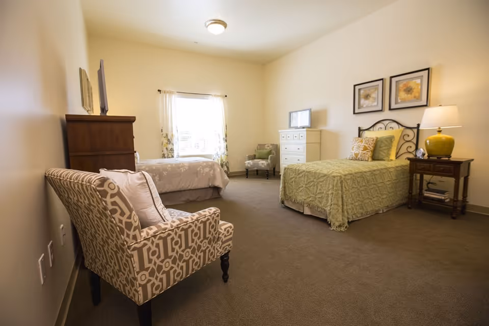 Spacious furnished bedroom with two twin beds, a patterned armchair, dresser, and a bedside table with lamp.