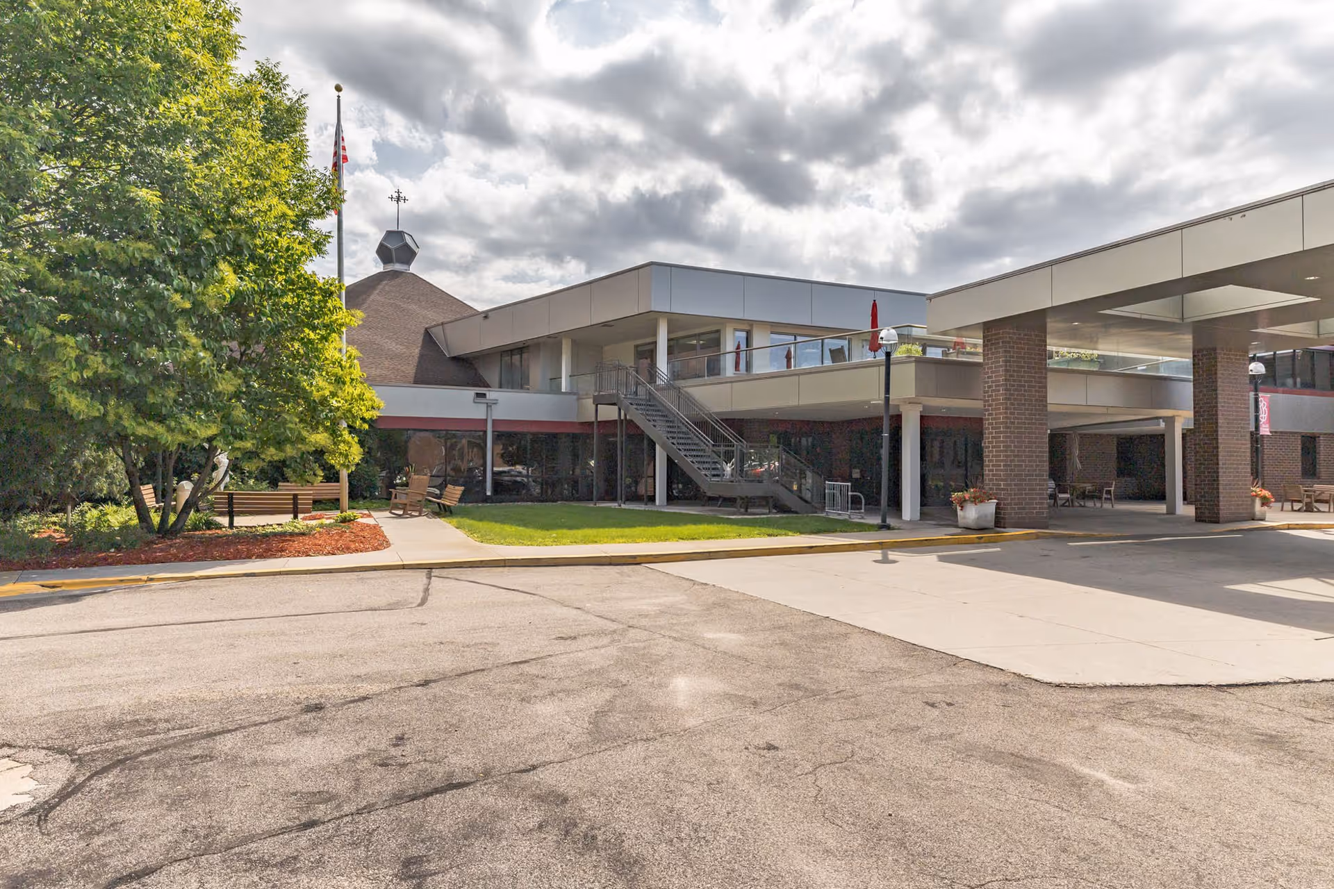 Exterior front entrance of a two-story senior living facility with a covered drop-off, outdoor seating, stairs and landscaping under a cloudy sky.