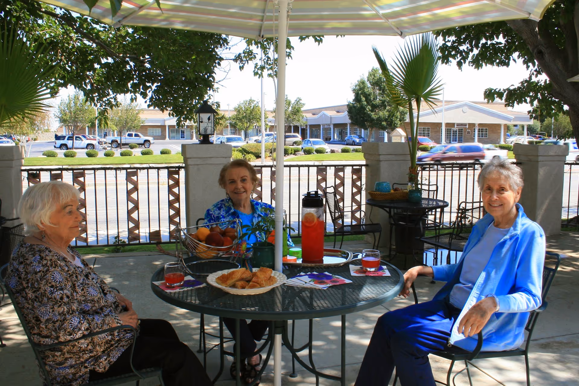 Three older women seated around a patio table under an umbrella enjoying drinks and snacks on an outdoor patio in front of a residential building.