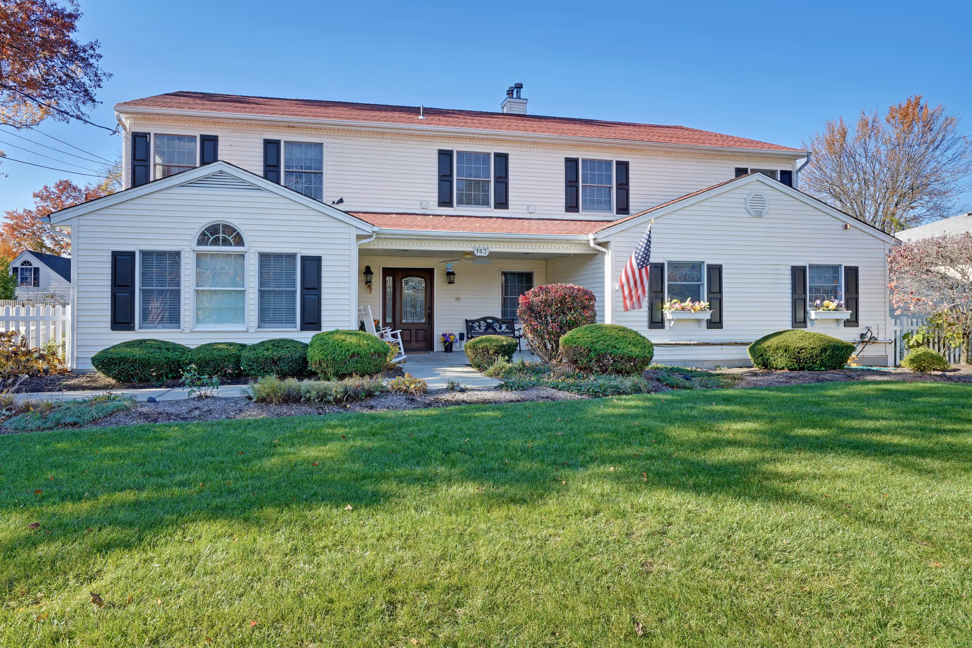 Front exterior view of a two-story white residential-style building with black shutters and a red roof, surrounded by neatly trimmed bushes and a green lawn. An American flag is displayed near the entrance, which has a covered porch with seating.