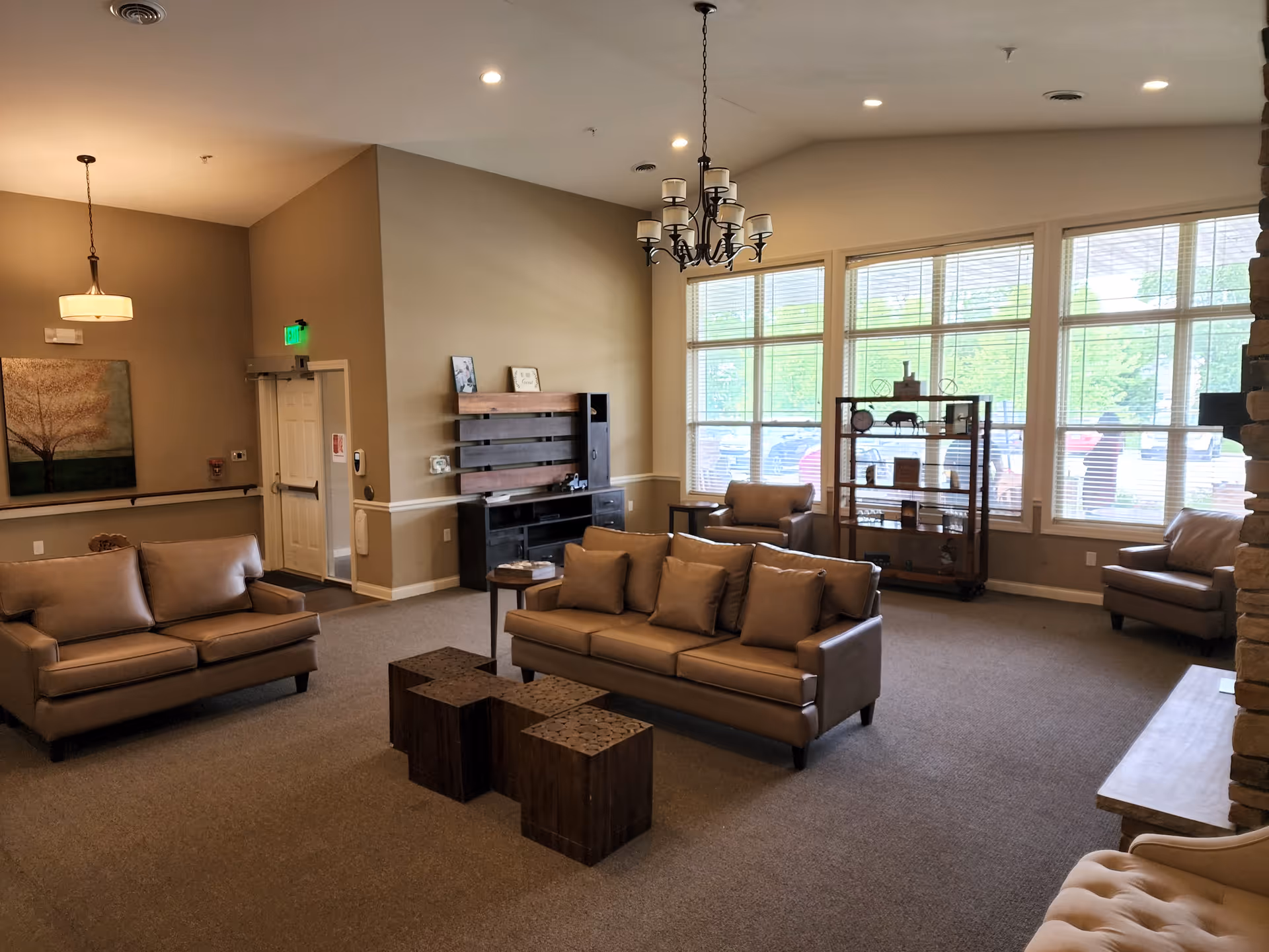 A spacious living room area with beige walls and carpeted floor, featuring two leather sofas with cushions, two armchairs, a wooden shelving unit with decorative items, a chandelier, and large windows letting in natural light.