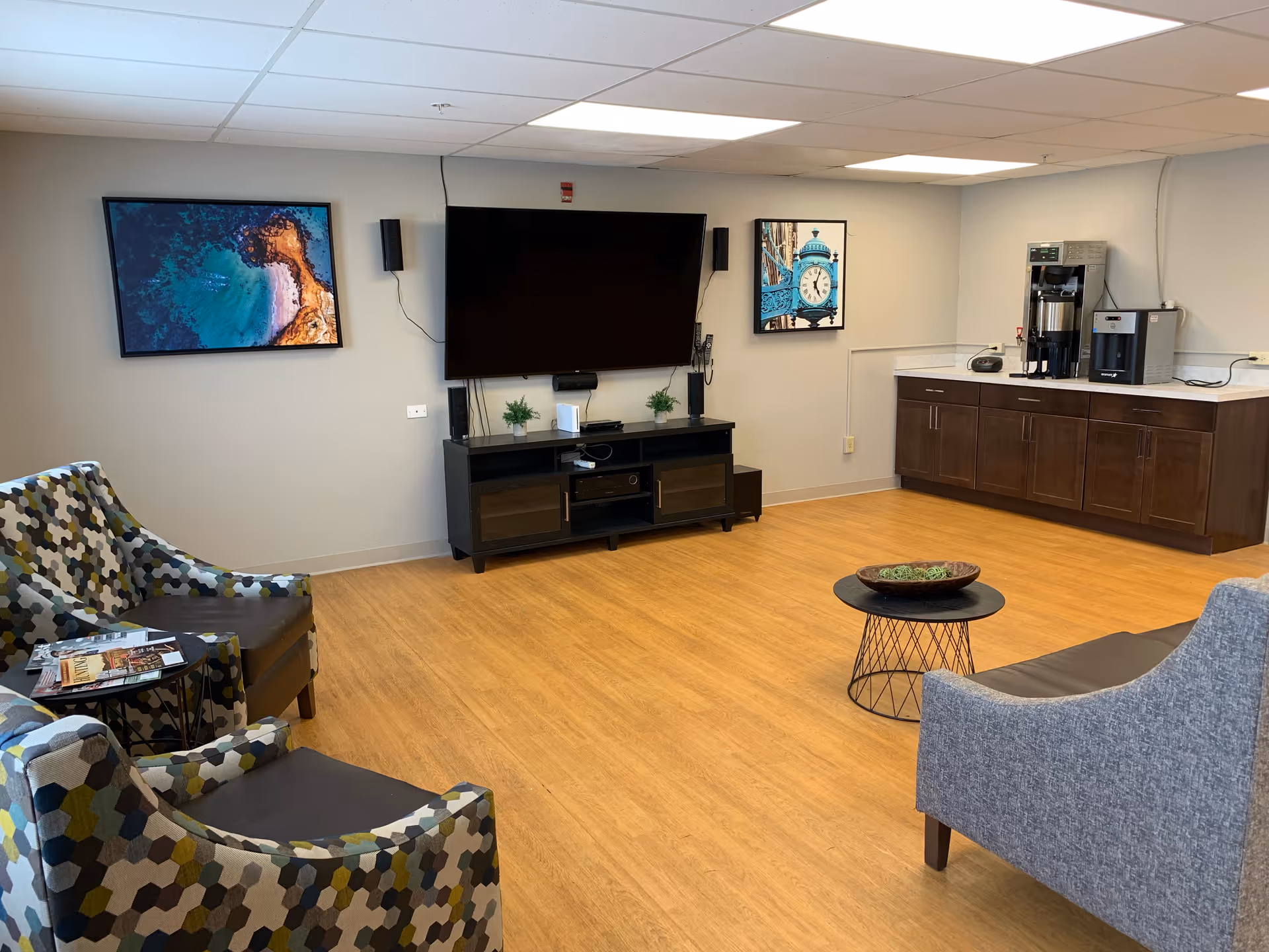 A comfortable common lounge with patterned armchairs, a large wall-mounted TV, a coffee station, and wood-look flooring.