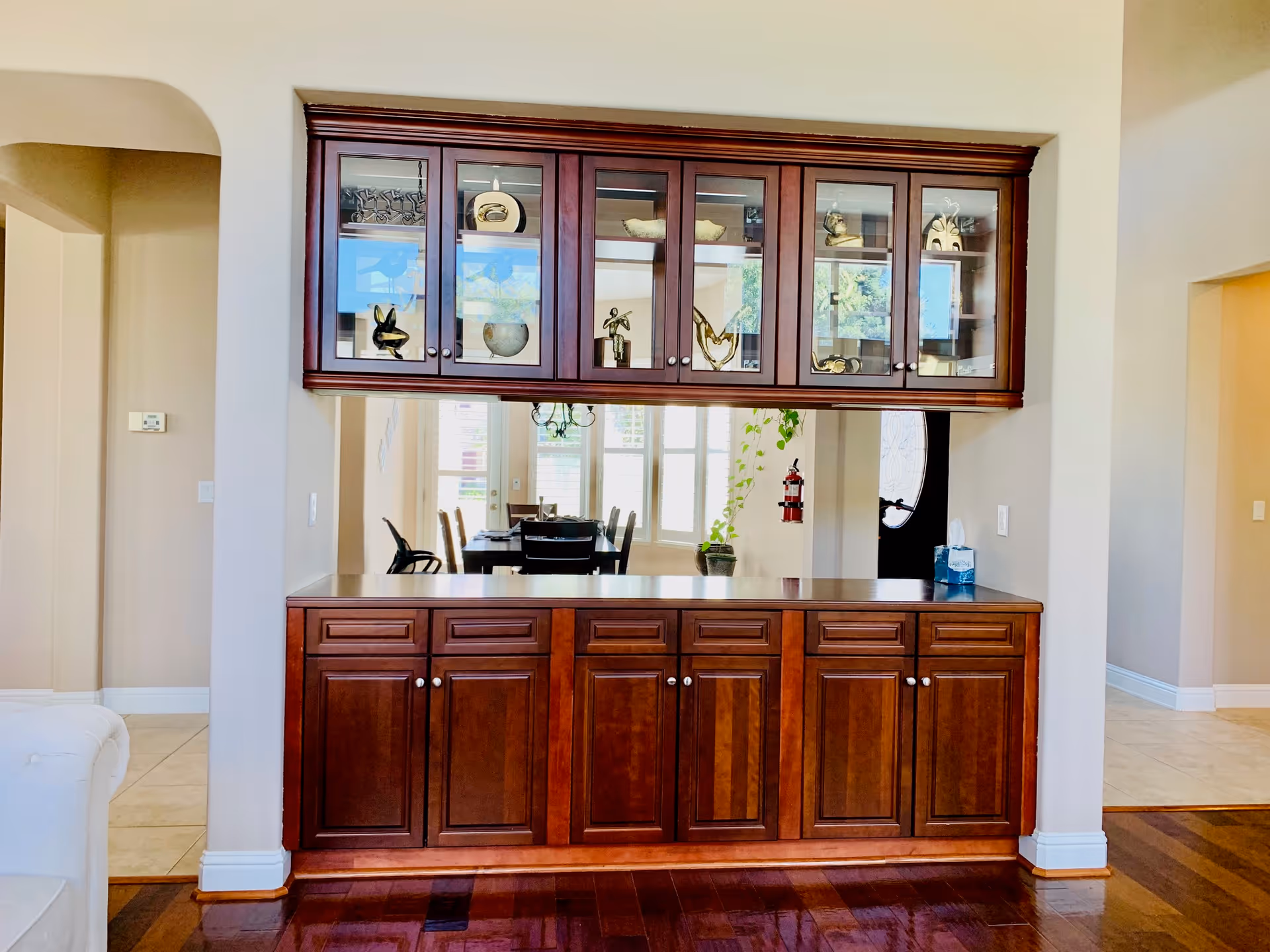 Interior view of a room featuring a wooden cabinet with glass doors on the upper section displaying decorative items, and solid wooden doors on the lower section. Behind the cabinet, a dining area with a table and chairs is visible through a large pass-through opening. The room has light-colored walls and a mix of tile and wood flooring.