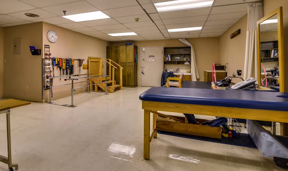 A physical therapy room with parallel bars, therapy steps, exercise belts hanging on the wall, a padded therapy table, a desk with a chair, and a door marked 'Not an Exit'. The room has beige walls and a tiled floor.