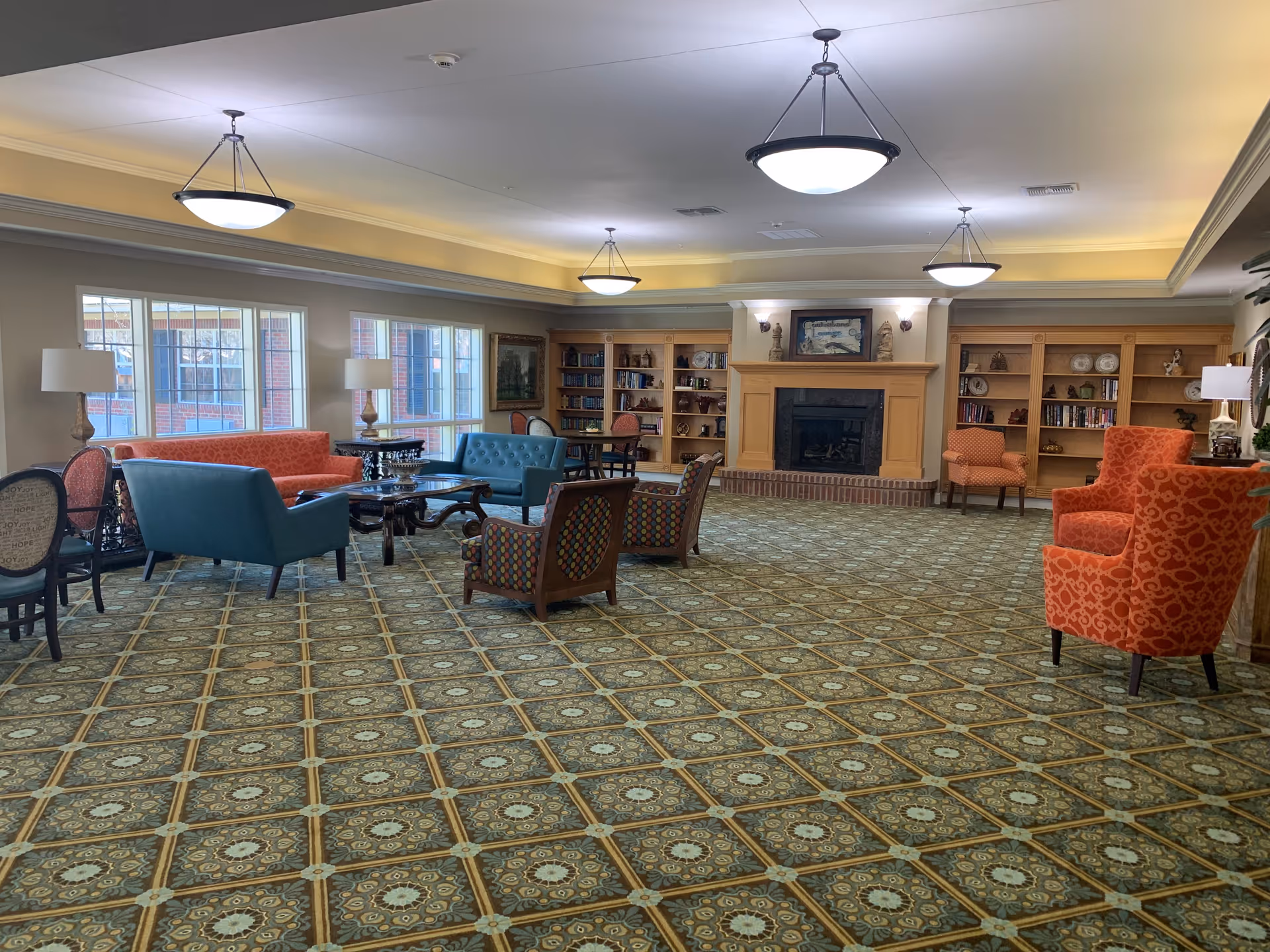 A spacious and well-lit common area with patterned carpet flooring, multiple seating arrangements including orange and blue upholstered chairs and sofas, wooden bookshelves filled with books and decorative items, a fireplace with a framed picture above it, and large windows letting in natural light.