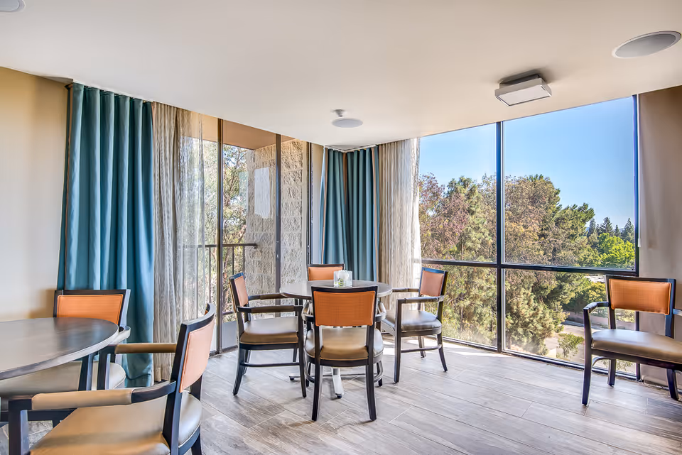 A bright dining area with round tables and chairs featuring orange backs and beige seats. Large floor-to-ceiling windows provide a view of green trees outside. Blue and beige curtains frame the windows, and the floor is covered with light-colored wood-like tiles.