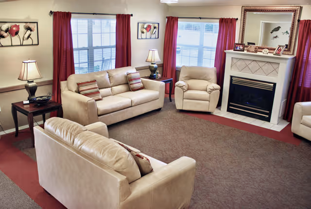 Well-lit living room with beige sofas and an armchair arranged around a fireplace, red curtains, and table lamps.