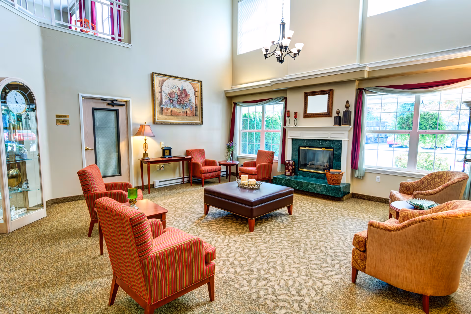 A bright and spacious living room with high ceilings, featuring a green marble fireplace, large windows with red curtains, and a seating area with striped and patterned armchairs arranged around a large brown ottoman. There is a wooden side table with a lamp and decorative items, a wall clock in a glass cabinet, and a floral painting on the wall.