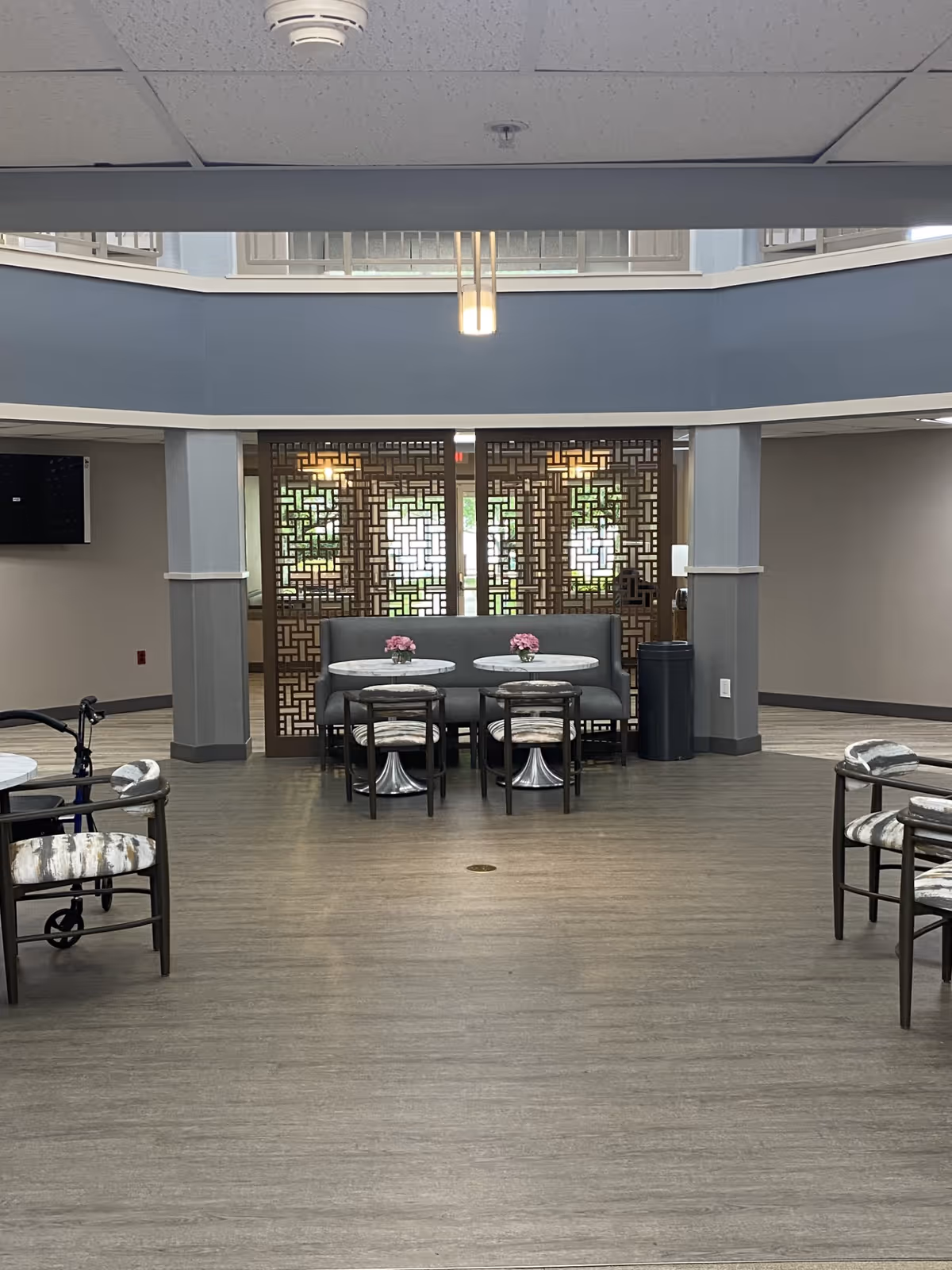 Interior common area with round tables and chairs arranged for seating. There is a gray cushioned bench against a decorative wooden partition with geometric patterns. The floor is wood-style vinyl, and the ceiling has recessed lighting and a hanging light fixture. A walker is visible to the left side of the image.