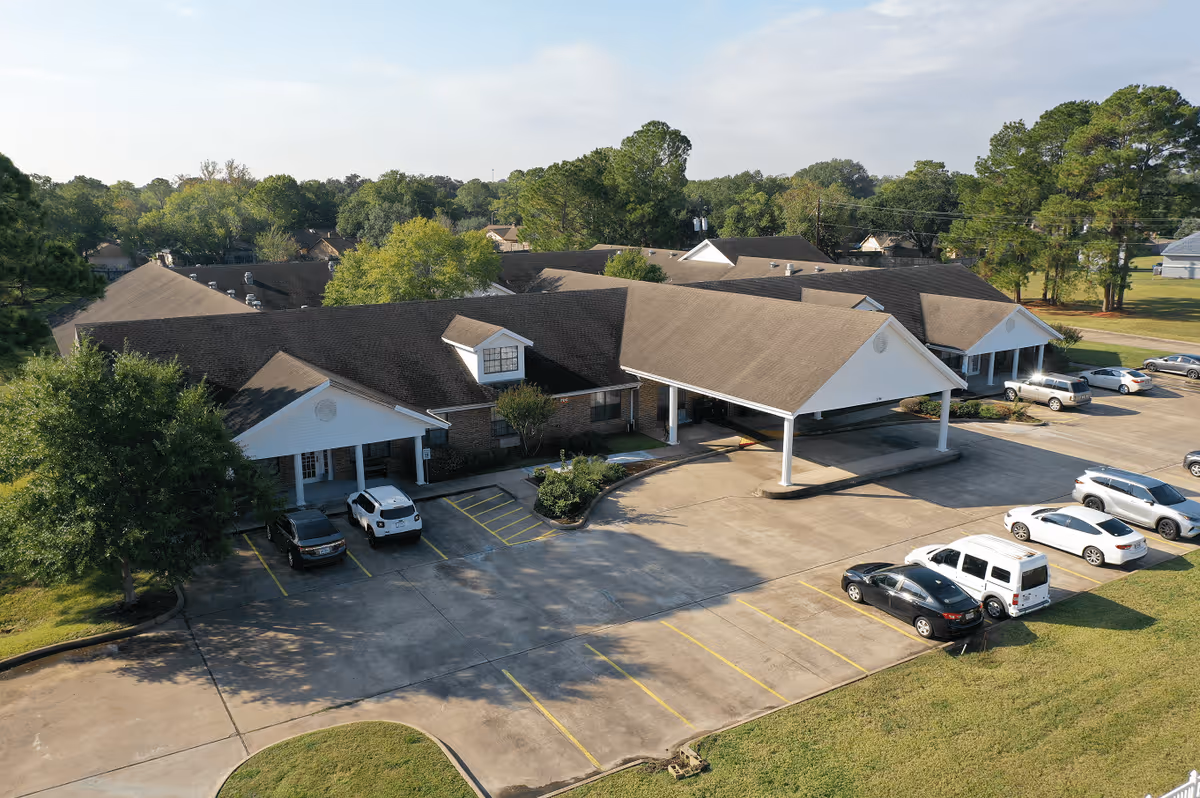 Aerial view of Cambridge Square Assisted Living facility showing a single-story building with a large covered entrance and several parked cars in the parking lot. The building is surrounded by trees and greenery under a partly cloudy sky.