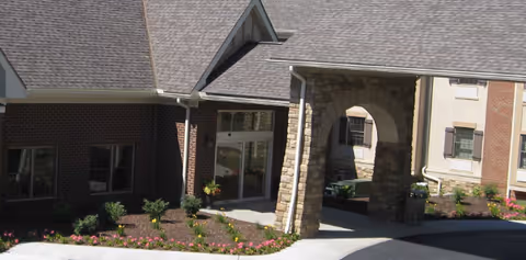 Exterior view of a senior living facility entrance with a covered stone archway, brick and beige walls, windows, and landscaped flower beds along the driveway.