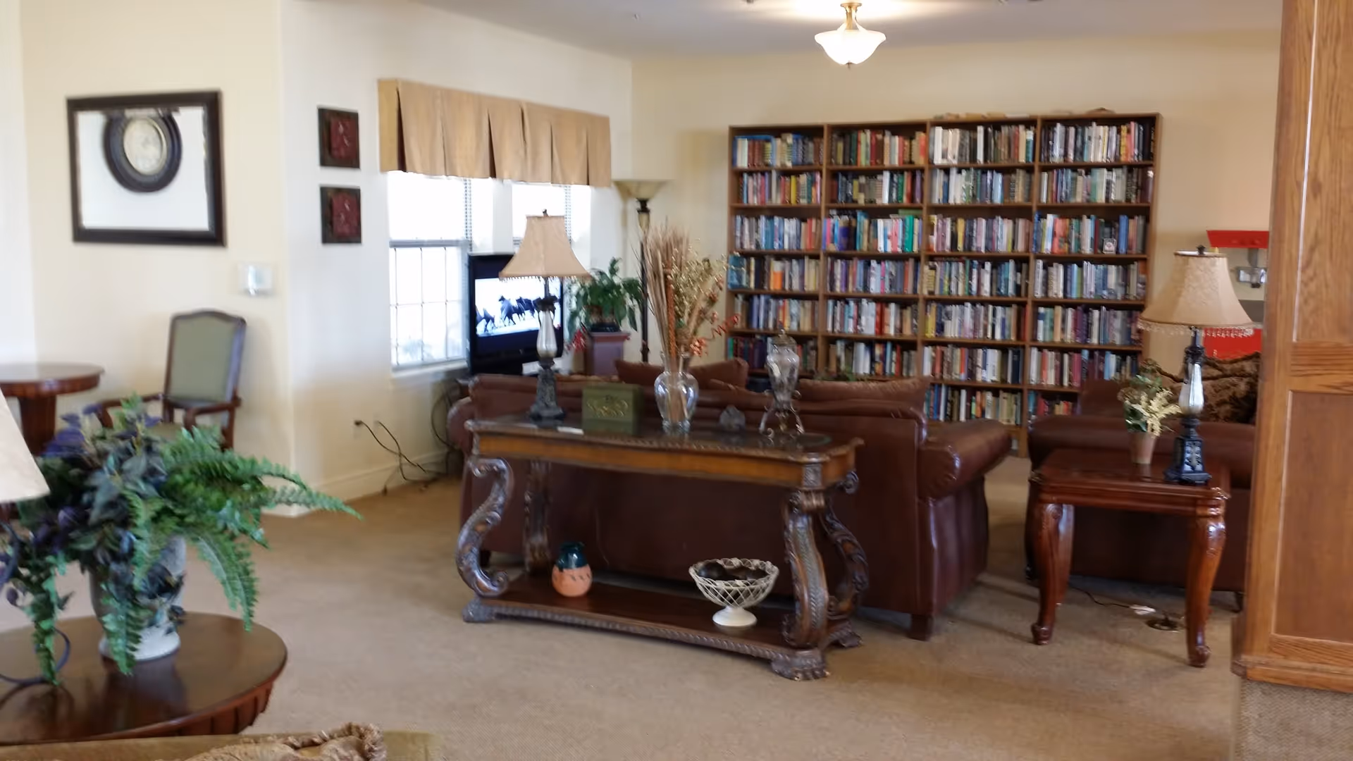 A cozy living room area with brown leather sofas, a large wooden bookshelf filled with books, two table lamps, and decorative plants. There is a TV near the window and a wooden console table with decorative items in the center of the room.
