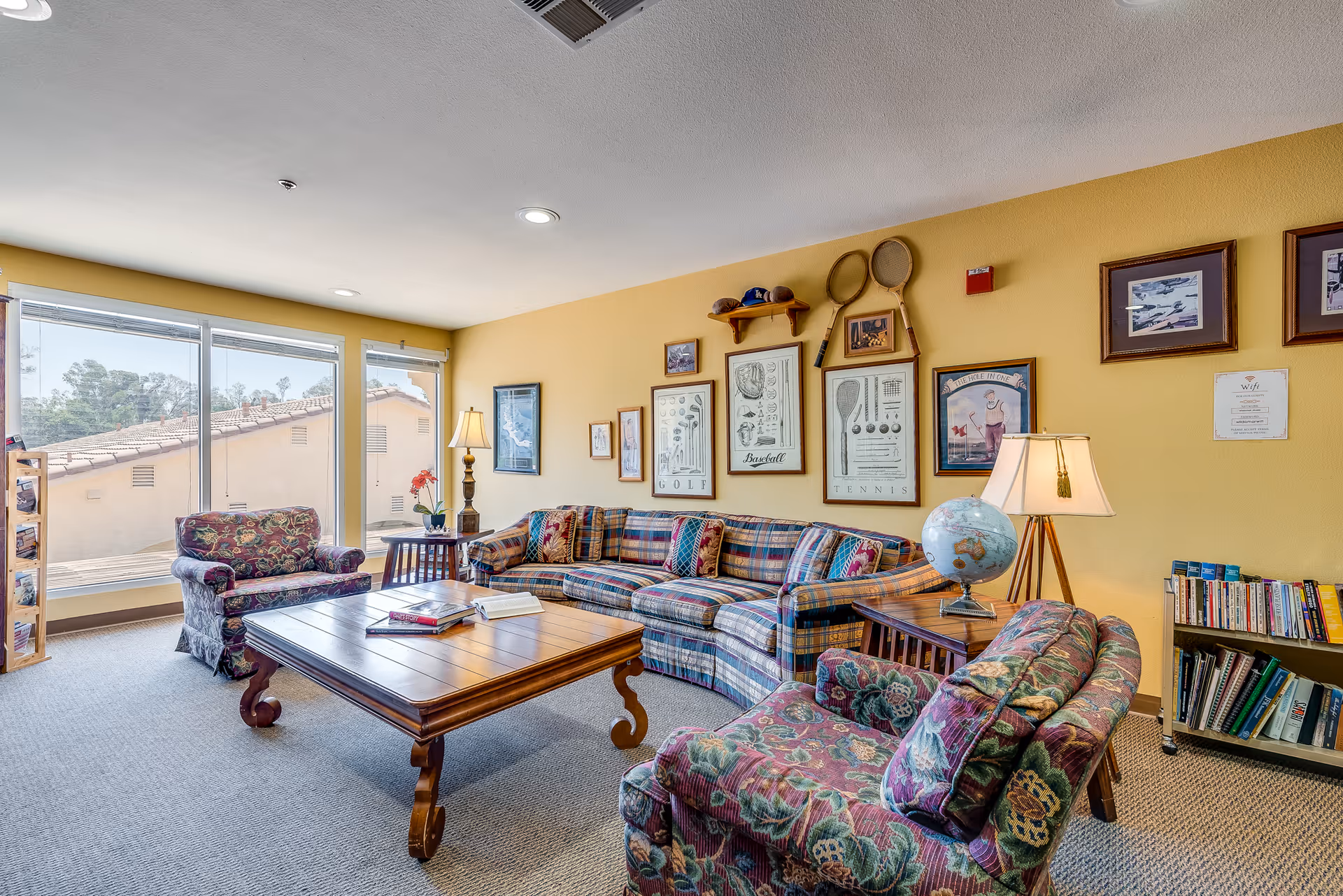 A cozy living room in a senior assisted living facility with large windows letting in natural light. The room features a plaid sofa, two floral upholstered armchairs, a wooden coffee table with books on it, and side tables with lamps. The walls are decorated with framed sports-themed artwork and vintage tennis rackets. A bookshelf filled with books is visible in the corner.