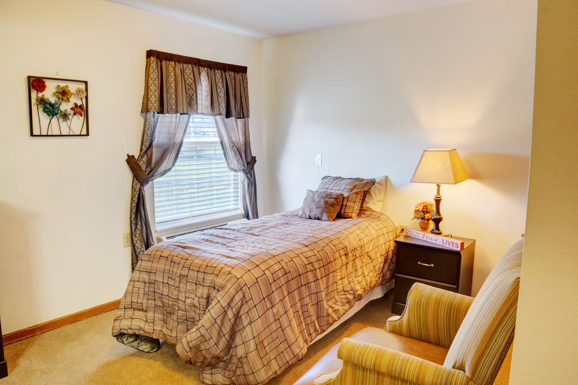 Small furnished bedroom with a twin bed, bedside table and lamp, armchair, and a curtained window.