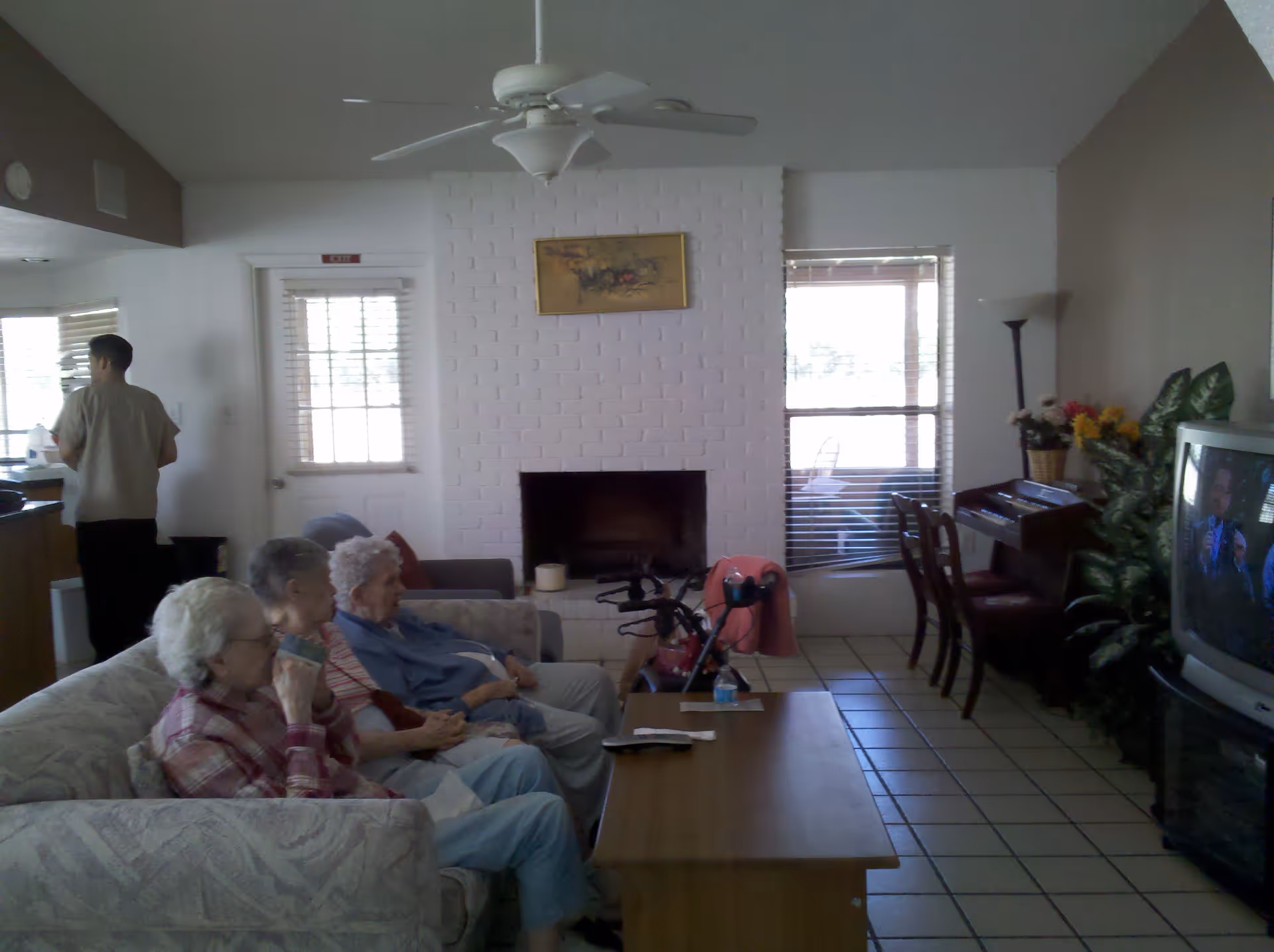 Three elderly women sitting on a couch watching television in a living room with a white brick fireplace, a ceiling fan, a wooden coffee table, and a man standing near the kitchen area in the background.