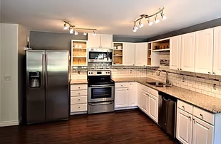 Modern kitchen with stainless steel refrigerator, oven, microwave, and dishwasher. White cabinets with granite countertops and subway tile backsplash. Dark wooden floor and ceiling track lighting.